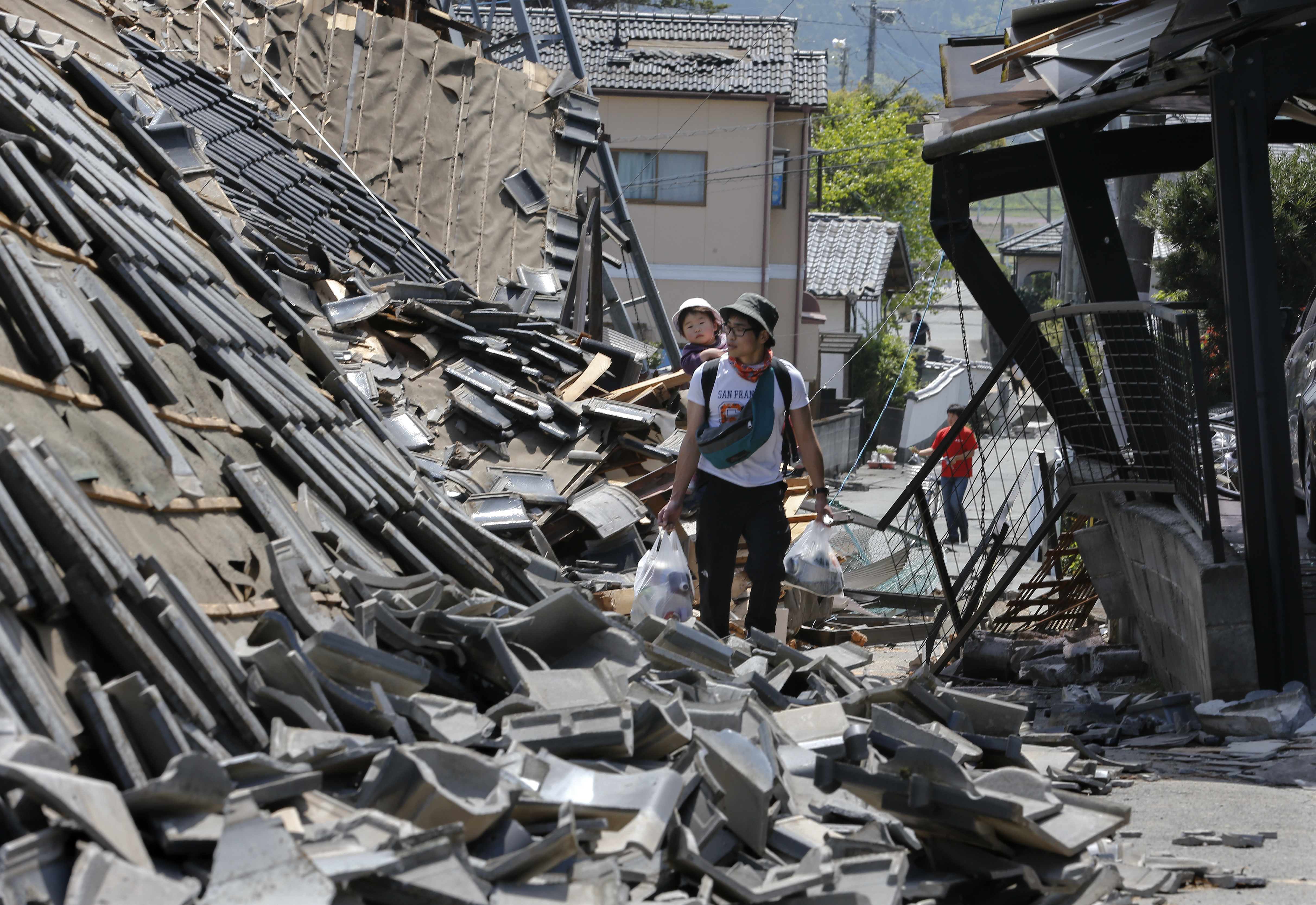 epa05259505 A resident carrying his child on his back walks through debris of collapsed houses with supplies in the town of Mashiki, where the strongest earthquake intensity of 7.0 on the Richter scale was registered for the first time since the 2011 T?hoku earthquake and tsunami, Kumamoto Prefecture, southwestern Japan, 15 April 2016. Strong earthquakes hit southwestern Japan the evening of 14 April. Authorities said nine people died and over 1,000 were injured by the earthquake, now named the Kumamoto Earthquake. Eight of the nine victims were reported in the town of Mashiki.  EPA/KIMIMASA MAYAMA